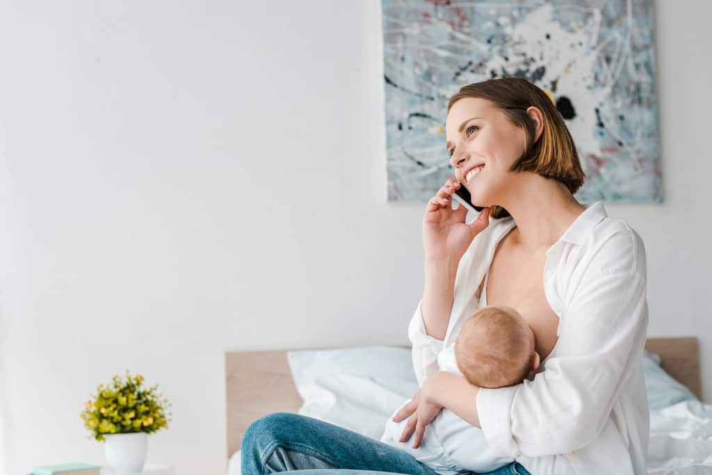 smiling woman talking on smartphone while breastfeeding baby at home